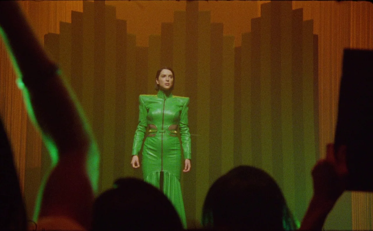 The Nowhere Inn (2020) movie still: medium — A woman stands on a dimly lit stage in front of a geometric backdrop. Out-of-focus silhouettes of…