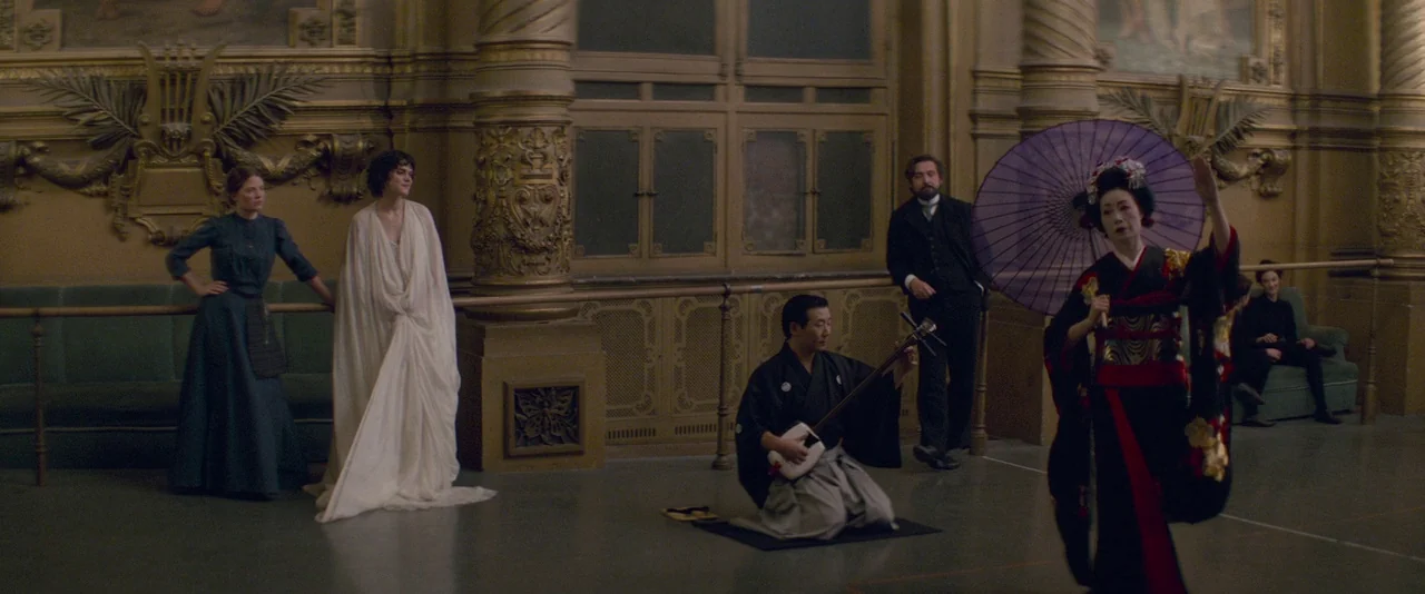 The Dancer movie still: wide — An opulent ballroom with ornate gold molding and spiraling columns features several individuals standing near walls.…