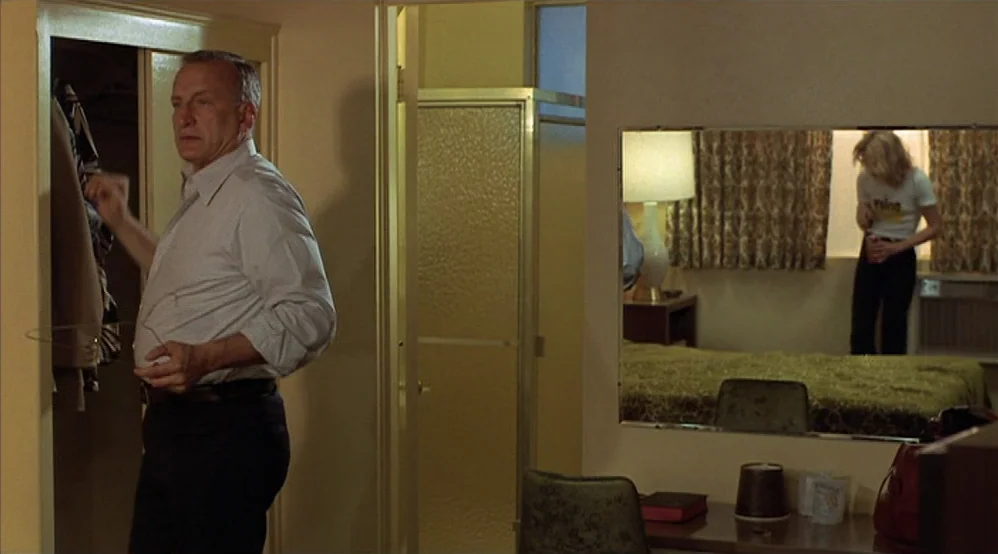 Hardcore (1979) movie still: medium — A standard motel interior features beige walls, a small vanity, and a large mirror. A man stands in the…