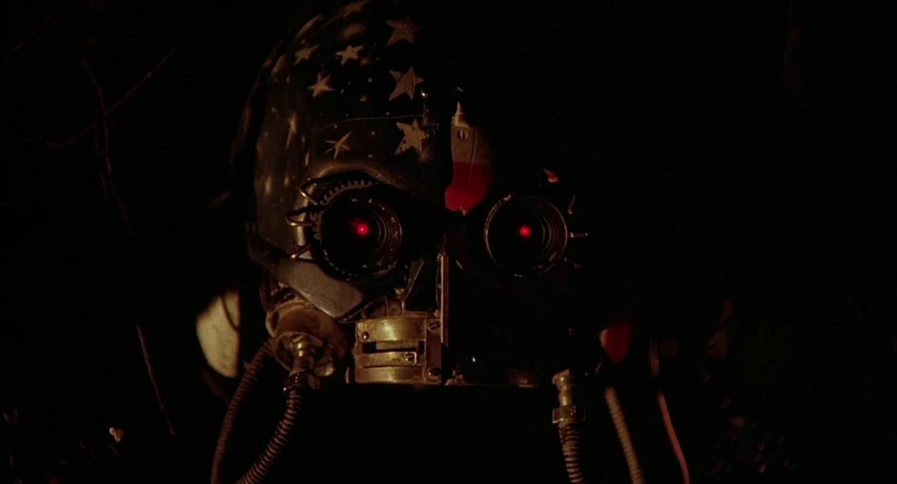 Hardware (1990) movie still: closeup — A dark space contains a metallic mask featuring white stars and glowing red lenses. The mask stares forward…