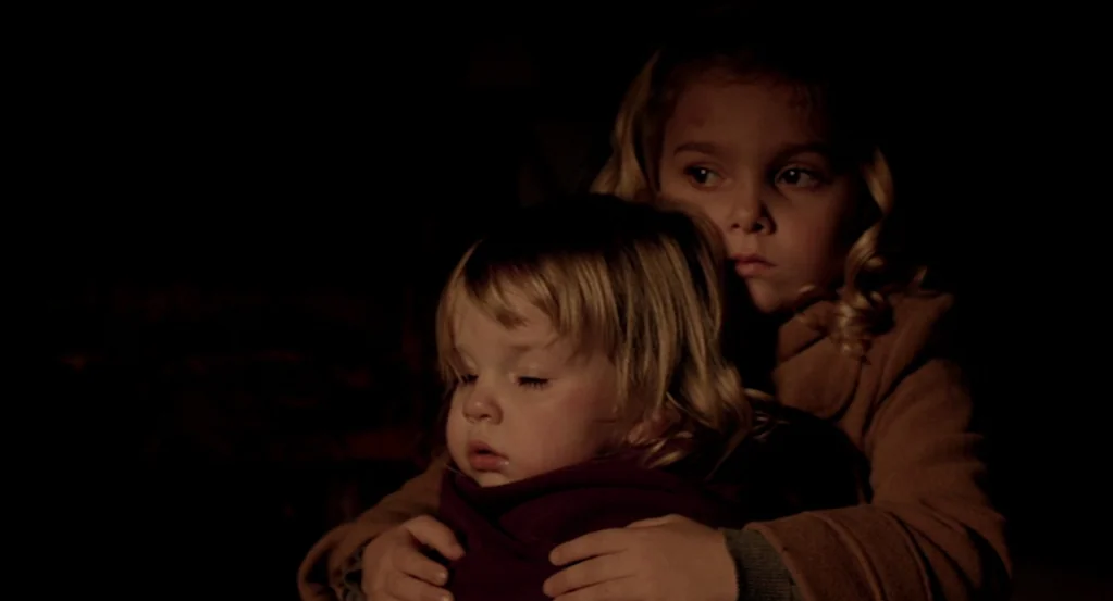 Mama (2013) movie still: medium — Two children sit together in a dark environment with very little visible surrounding detail. The older child holds…
