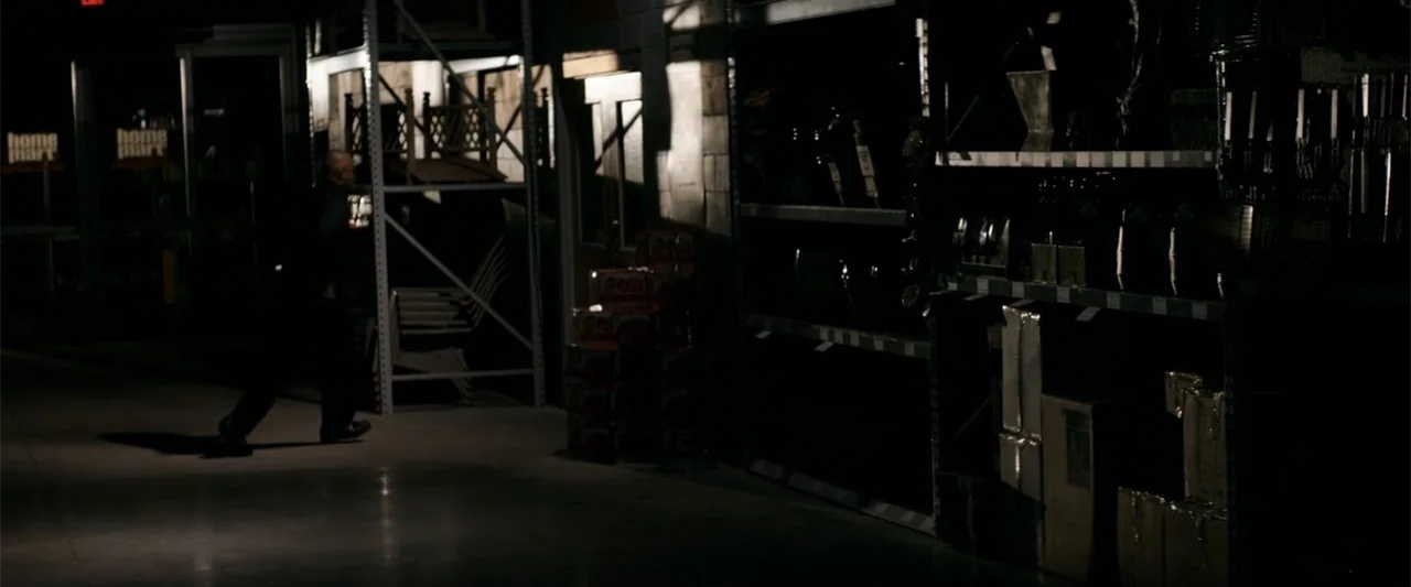The Equalizer (2014) movie still: wide — A dark retail store aisle is filled with tall industrial metal storage shelves. A man walks through the dim…