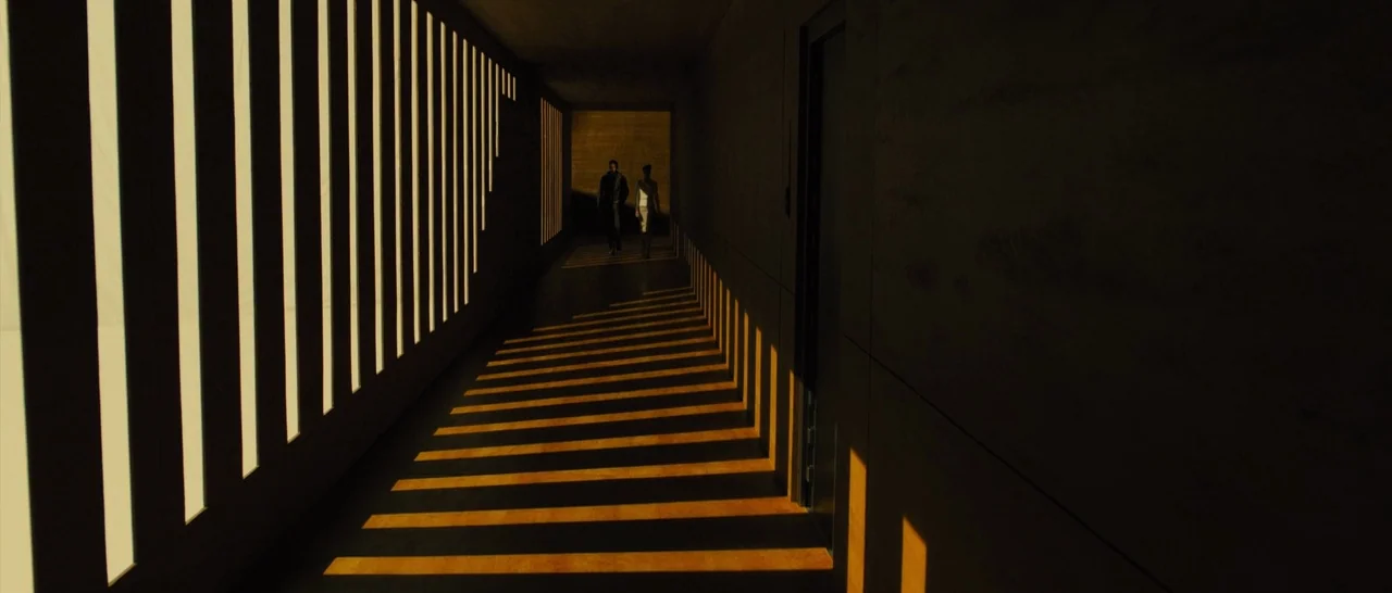 Blade Runner 2049 (2017) movie still: wide — A long, minimalist concrete hallway features floor-to-ceiling vertical slats along one wall. Two…