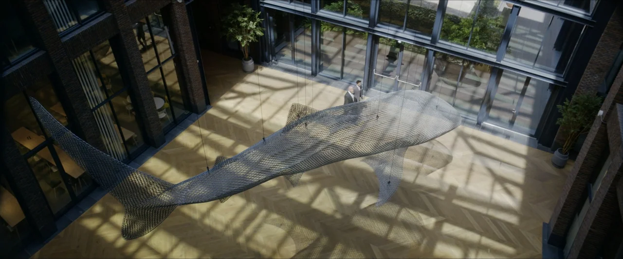 Black Lotus (2023) movie still: wide — A spacious office atrium features a large wire sculpture shaped like a whale. Two individuals walk across the…
