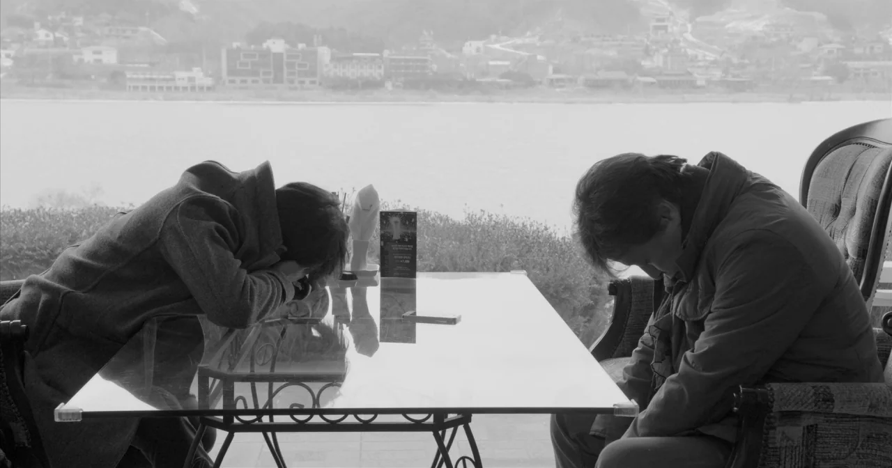 Hotel By The River movie still: medium — A glass table sits in front of a wide window overlooking a river landscape. Two men sit opposite each other…