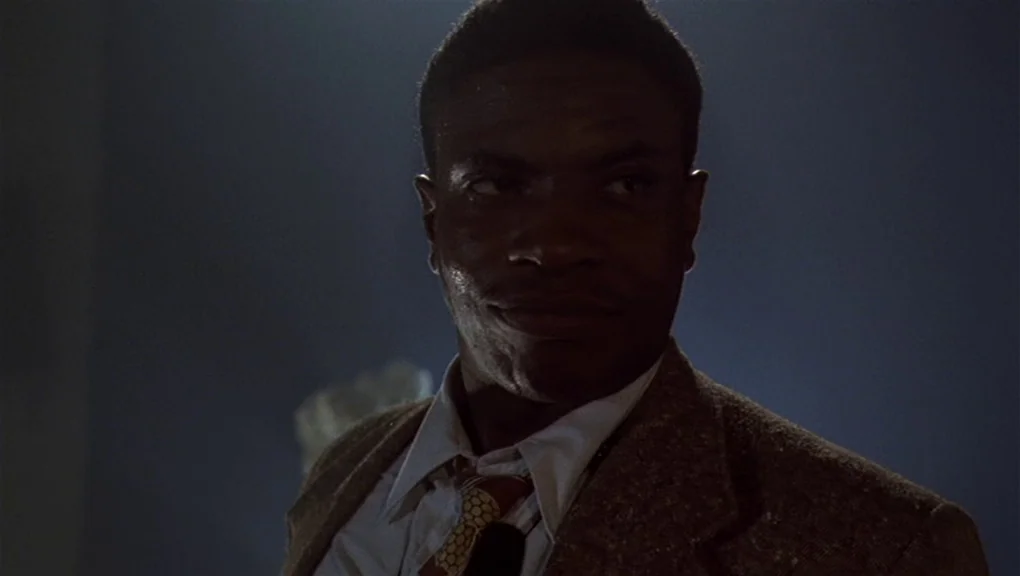 Bird (1988) movie still: closeup — A dimly lit interior space with shadows cast across the back wall. A man in a blazer turns his head and looks…