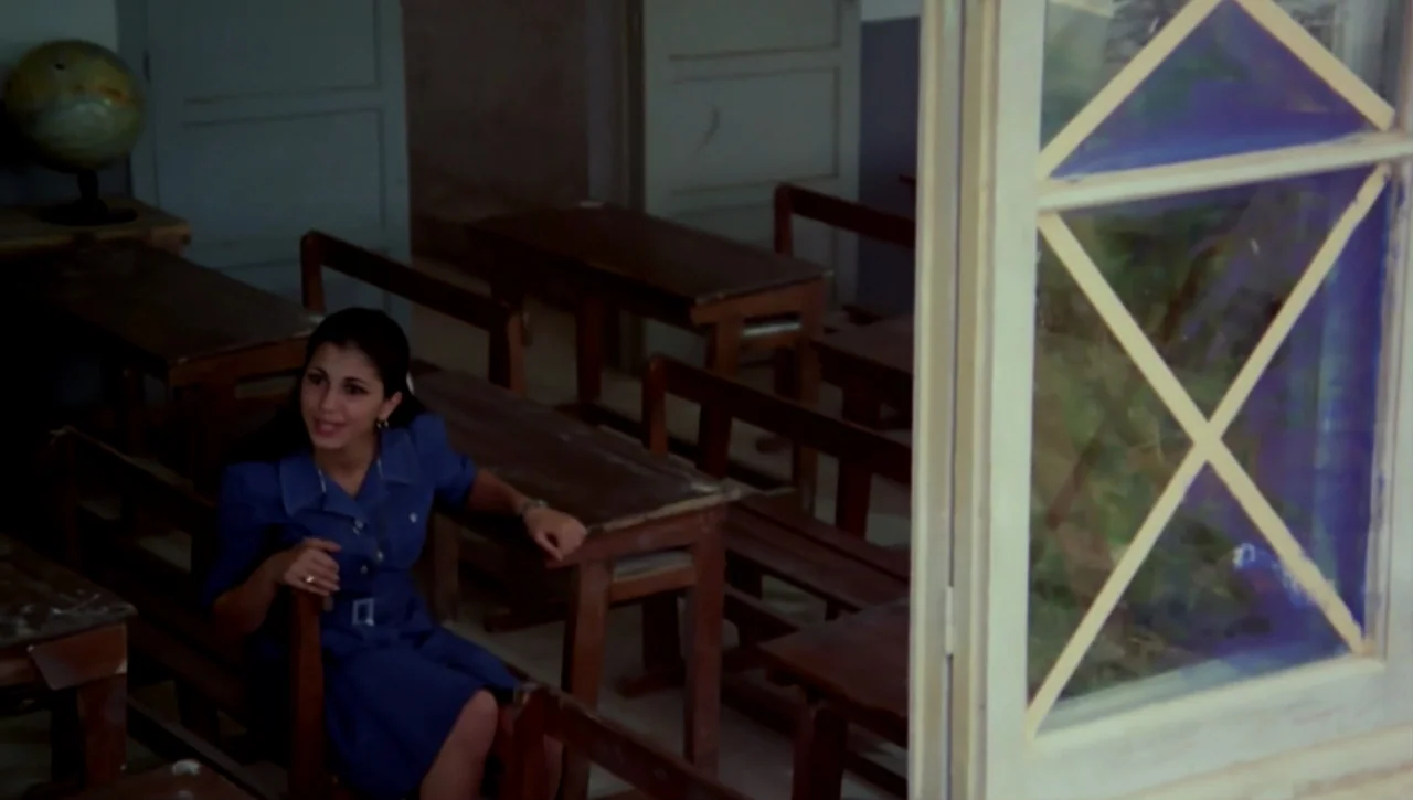 The Return of the Prodigal Son (1978) movie still: medium — A sparsely furnished room contains rows of simple wooden student benches and desks. A…