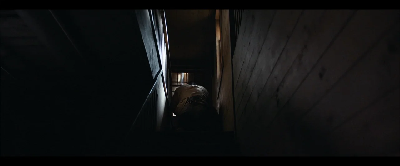It Comes at Night (2017) movie still: medium — A narrow, dark wooden staircase interior features horizontal wall planks and a single window. A man…