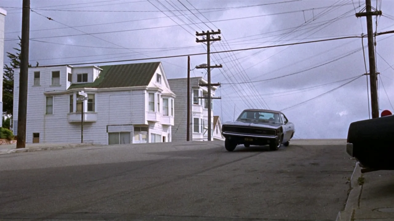 Bullitt (1968) movie still: wide — A residential San Francisco street features white houses and many overhead power lines. A black muscle car drives…