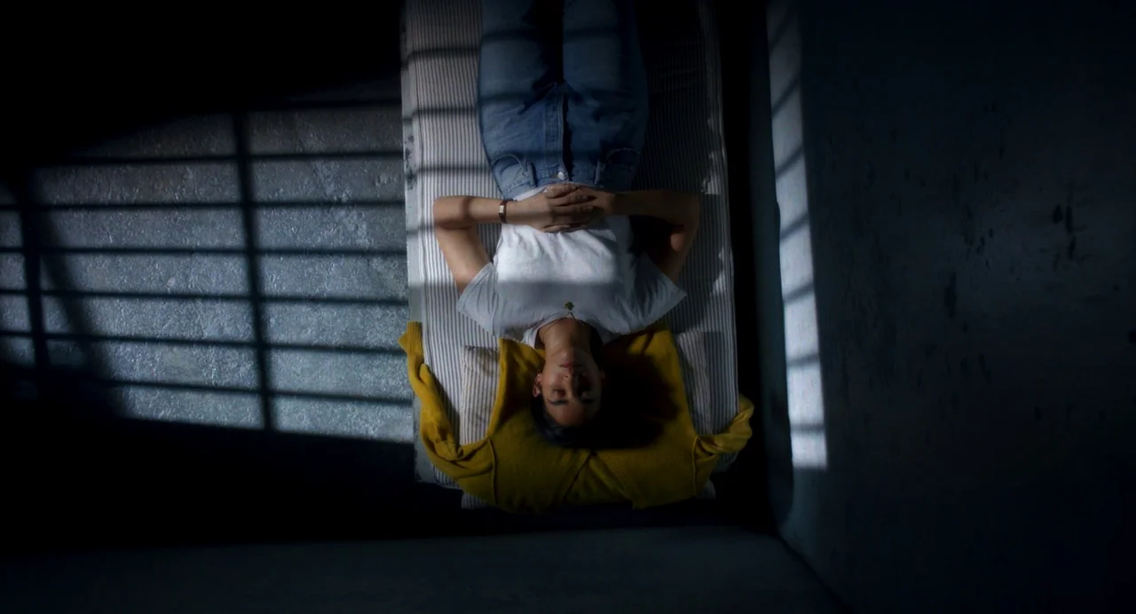 Drive-Away Dykes (2024) movie still: fullbody — A stark room with bare concrete walls and a narrow striped mattress on floor. A person lies flat on…