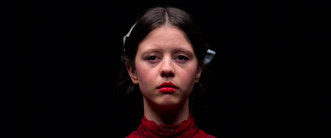 Pearl (2022) movie still: closeup — A woman stands centered against a completely black background. She faces forward with a direct, steady gaze.