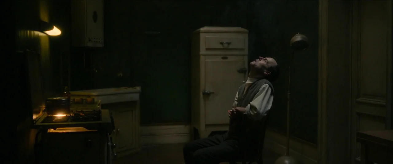 Earwig (2022) movie still: wide — A dimly lit 1940s kitchen contains a vintage stove and a tall white refrigerator. A lone man sits on a chair in…
