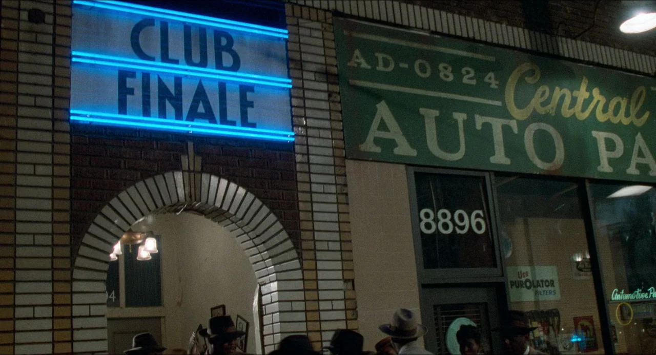 Devil In A Blue Dress (1995) movie still: establishing — A night view of a brick building featuring a blue neon club sign. Adjacent is an auto parts…
