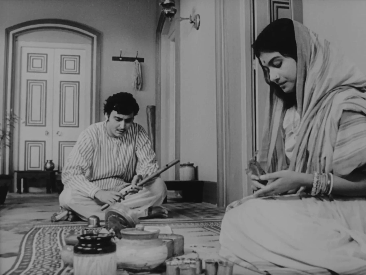 Charulata (1964) movie still: medium — An interior space features traditional patterned flooring and a central doorway in the back. A young man sits…