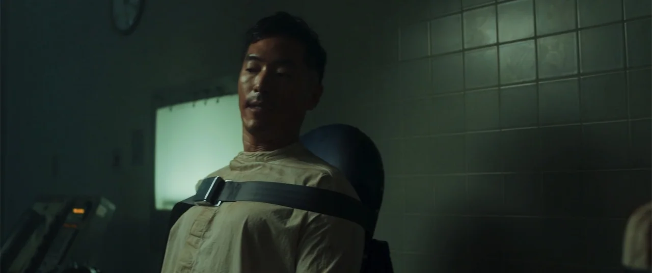 Phobias (2021) movie still: medium — A dimly lit hospital room features tiled walls and a bright light panel behind. An East Asian man sits in a…