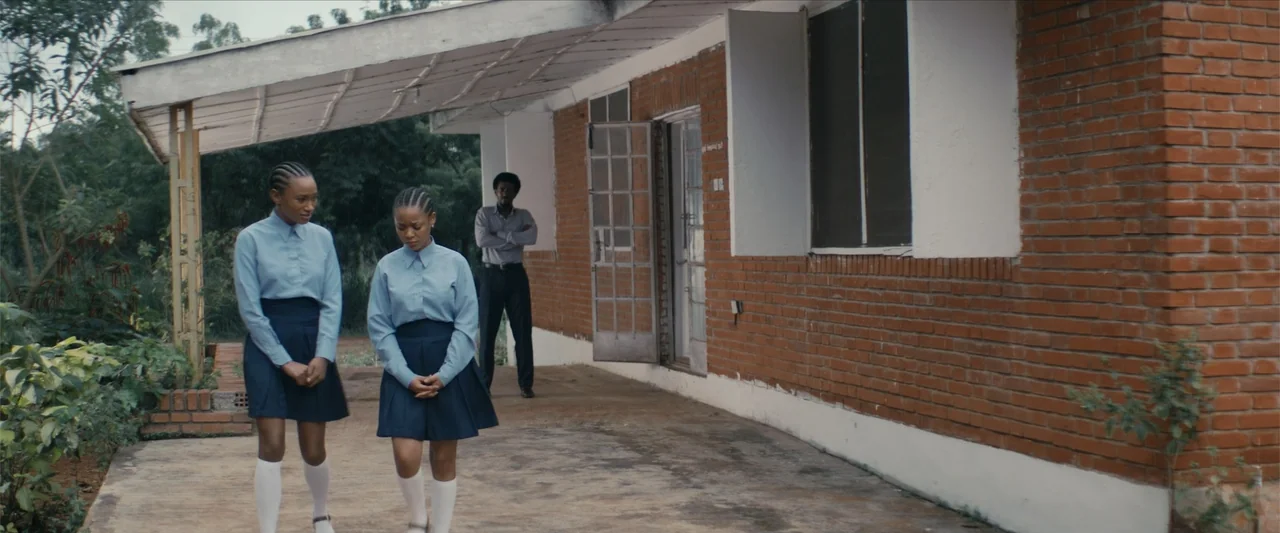 Ms. Kanyin (2025) movie still: fullbody — A brick building with a concrete walkway and a covered exterior porch area. Two teenage girls in school…