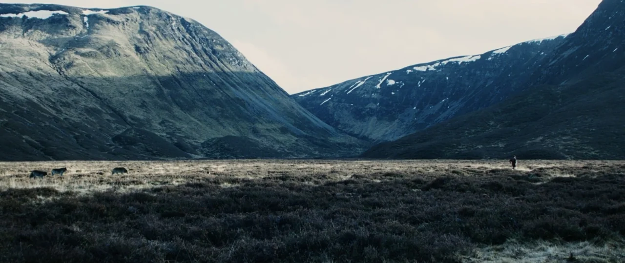 Centurion (2010) movie still: establishing — A vast, open moorland sits beneath steep, dark mountains with patches of snow. A small human figure…