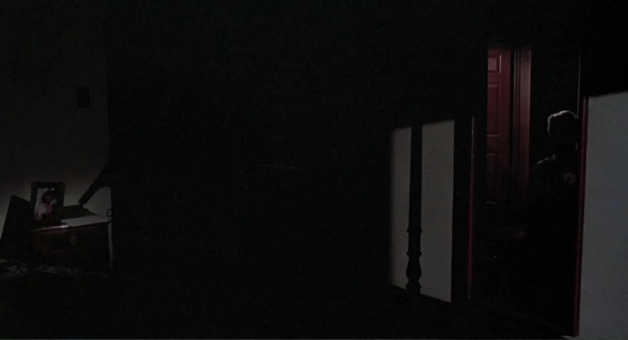 Dead of Winter (2025) movie still: wide — A dark bedroom features a small illuminated picture frame on a wooden side table. A person is silhouetted…