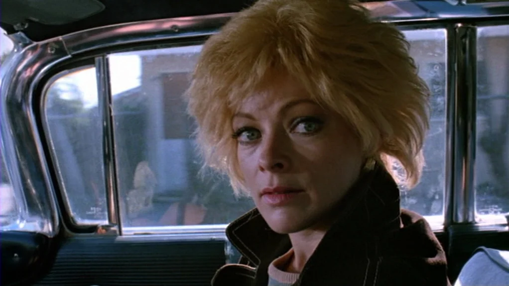 Patty Hearst (1988) movie still: closeup — The interior of a parked car with metal window frames and blurred suburban surroundings. A blonde woman…