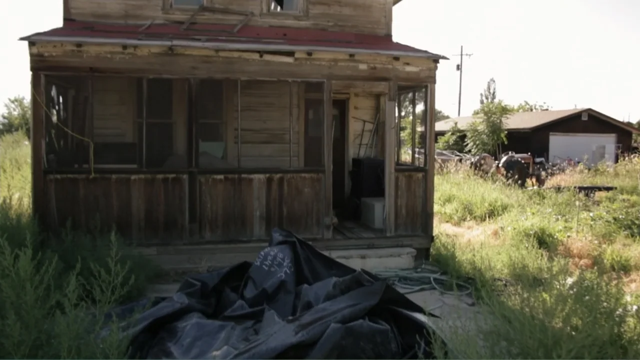 Savageland (2015) movie still: fullbody — A weather-beaten wooden house features a covered porch and a red metal roof. A pile of black plastic…