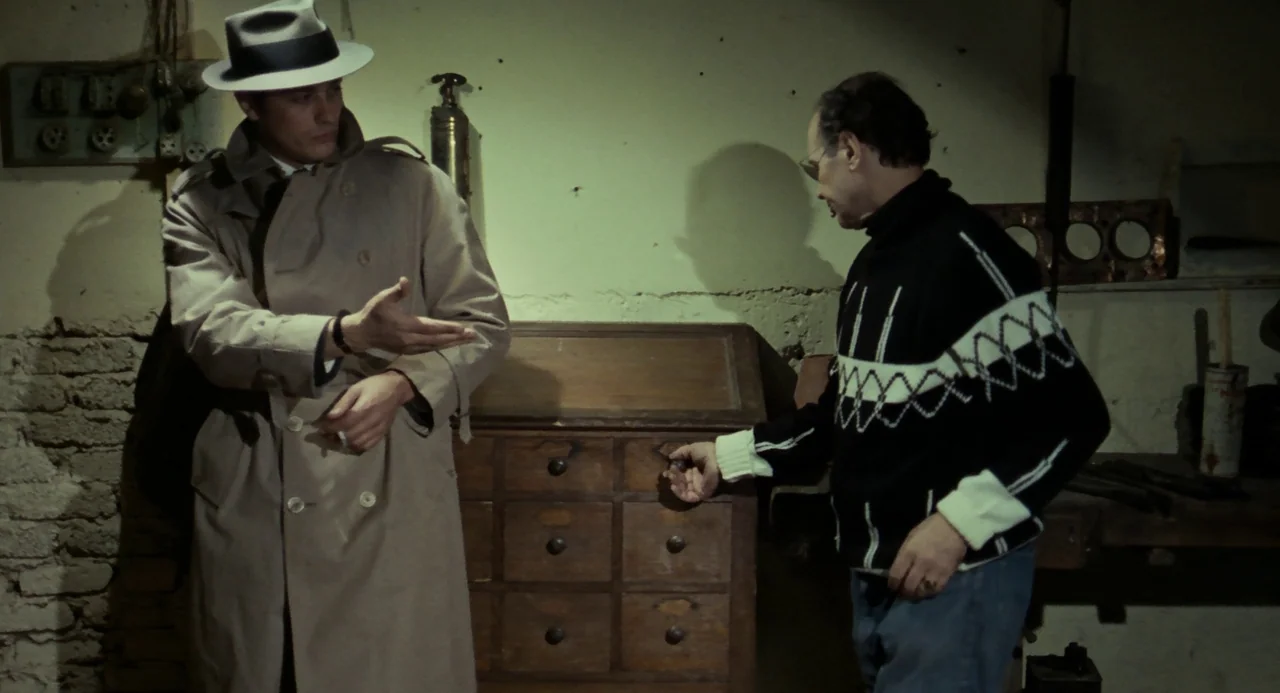 Le Samouraï (1967) movie still: medium — A basement room with bare stone walls and a small wooden storage cabinet. One man in a trench coat observes…
