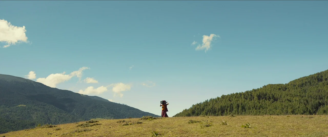 The Monk and The Gun (2023) movie still: wide — A vast, open hillside covered in short, patchy grass overlooks a distant pine-covered mountain. A…