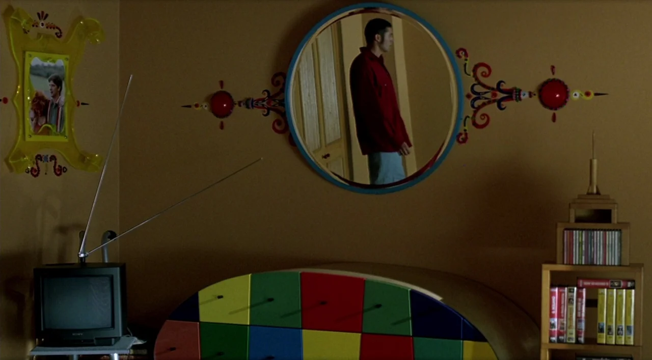 Kika (1993) movie still: medium — A wall in a room features a circular mirror and colorful patterned decorations. A young man is visible standing…