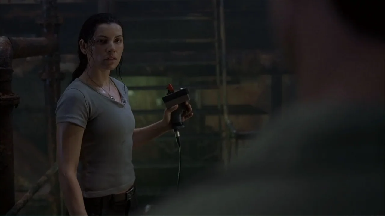 Ghost Ship (2002) movie still: medium — A dark industrial setting features metal staircases and piping in the blurred background. A woman in a grey…