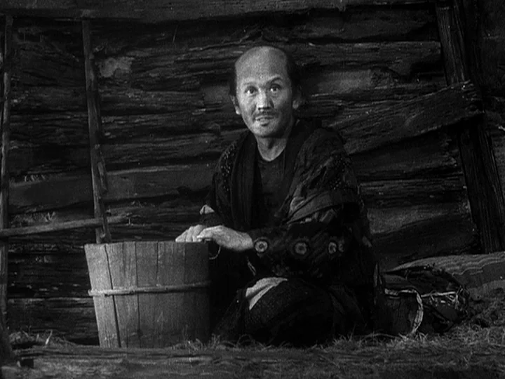 The Lower Depths (1957) movie still: medium — A rustic shack features walls constructed from dark, rough-hewn horizontal timber logs. A man with a…