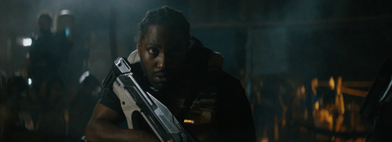 The Creator (2023) movie still: medium — A dimly lit industrial setting with indistinct machinery and hardware visible in the background. A man…
