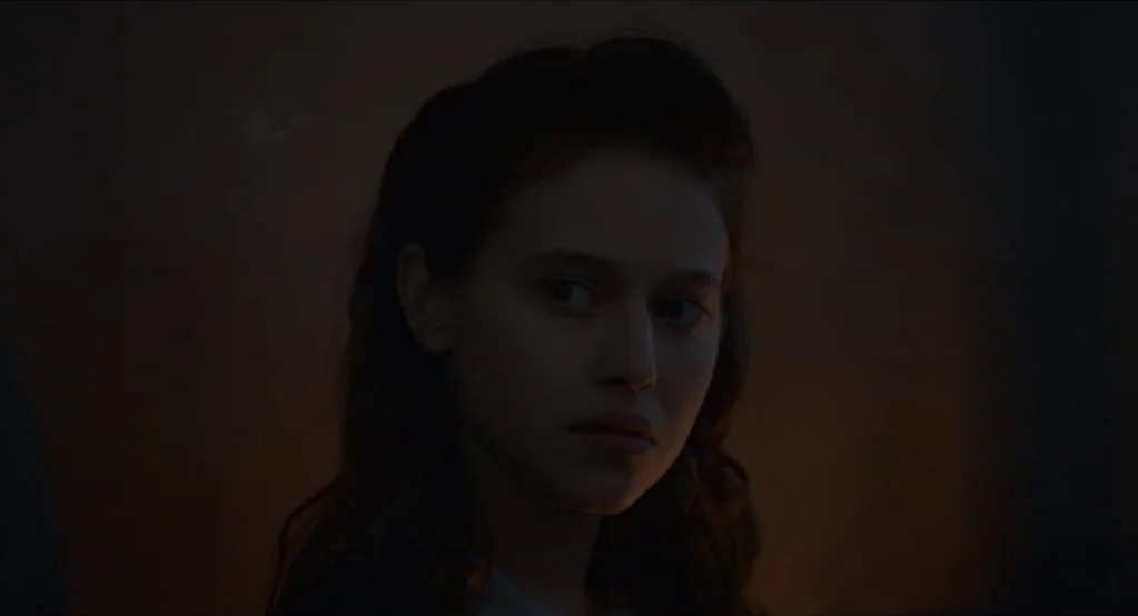 The Innocents (2016) movie still: closeup — A dark indoor setting with low ambient light from an off-screen source. A young woman with dark hair…
