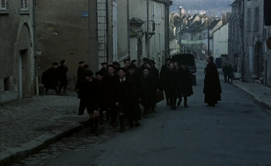 Au Revoir Les Enfants (1987) movie still: wide — A narrow cobblestone European street is flanked by tall, weathered stone residential buildings. A…