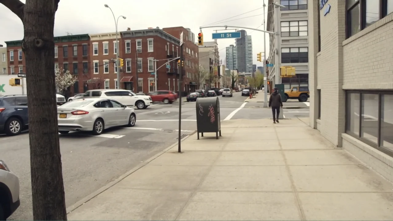 Suited (2016) movie still: wide — An urban intersection with brick buildings and a metal mailbox on a concrete sidewalk. A person walks along the…