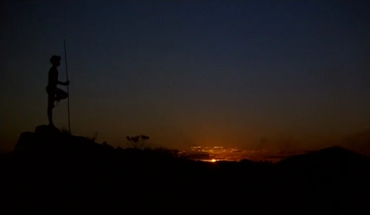 Walkabout (1971) movie still: wide — A dark rocky hilltop overlooks a desert landscape during the final moments of sunset. A lone figure stands on…