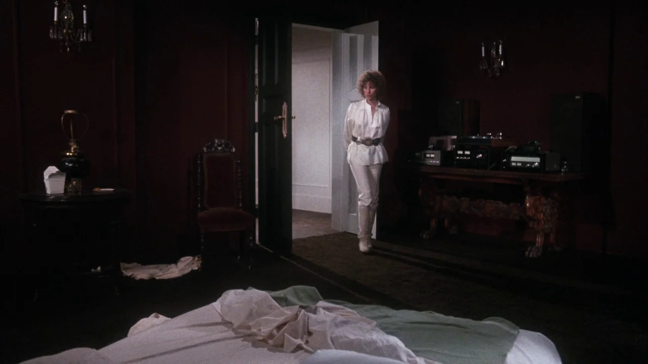 A Star Is Born (1976) movie still: fullbody — A dimly lit room with dark walls, antique furniture, and a messy bed in foreground. A woman in white…