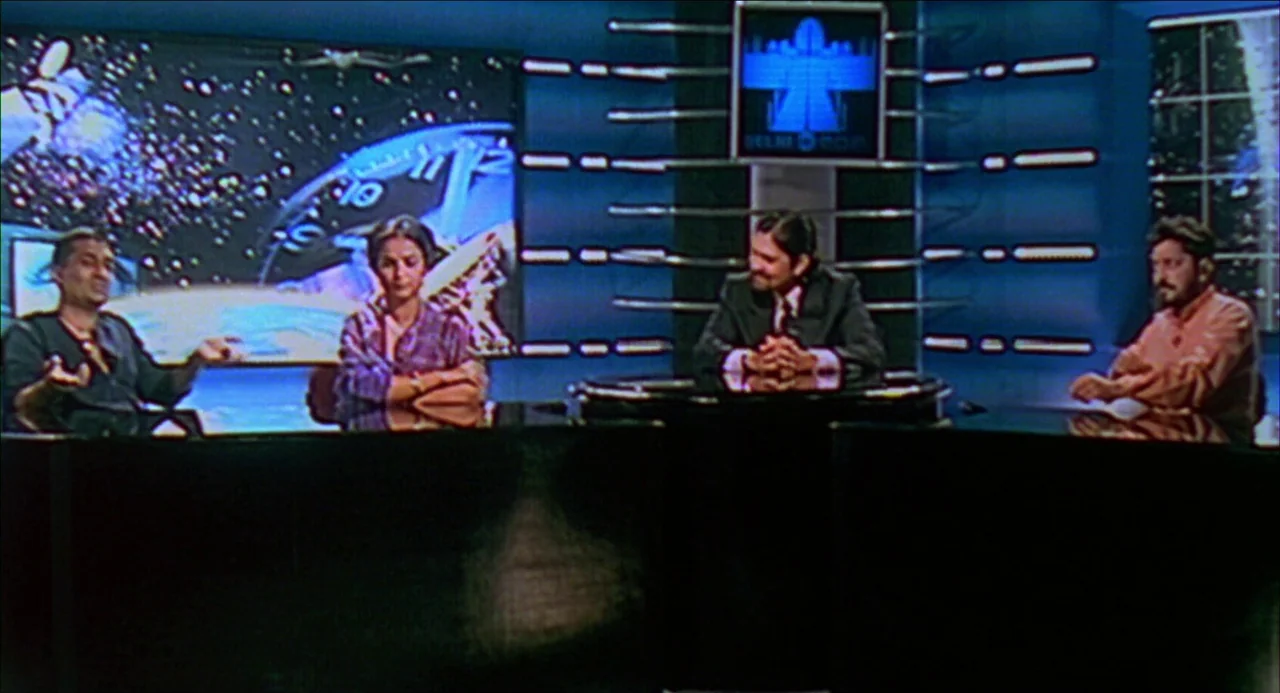 Monsoon Wedding (2001) movie still: wide — A television broadcast set features a curved desk and large screens showing space imagery. Four people…