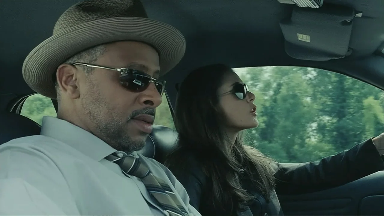 Mr. Brooks (2007) movie still: medium — Inside a moving car, a man in a hat and a woman drive along a road. The woman behind the wheel has her mouth…