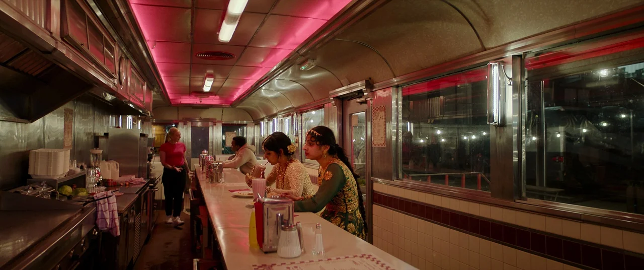 Polite Society (2023) movie still: wide — A classic stainless-steel interior diner features a long white counter and booth seating. Two young women…