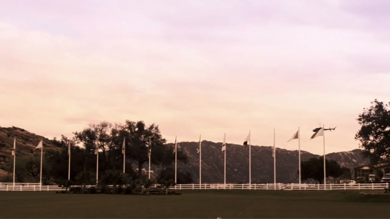 Redline (2007) movie still: establishing — A grassy field is bordered by a white fence and a row of tall flagpoles. A distant helicopter flies over…