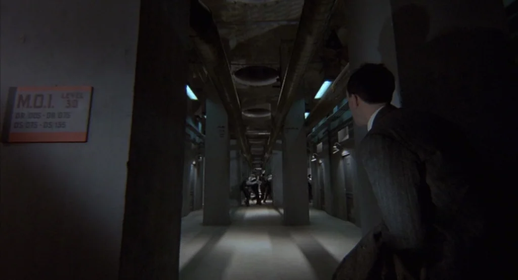 Brazil (1985) movie still: medium — A long industrial corridor features concrete pillars and multiple metal doors on both sides. A man in a suit…