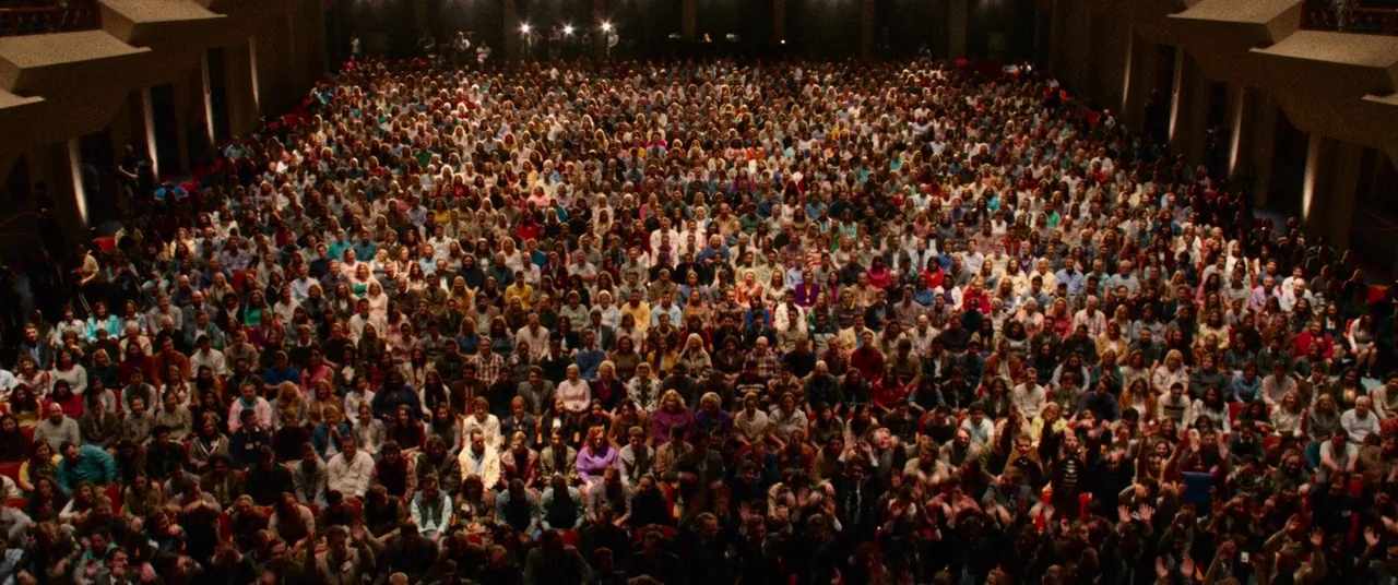Steve Jobs (2015) movie still: establishing — A massive indoor auditorium is filled with hundreds of people seated in rows. The crowd is applauding…