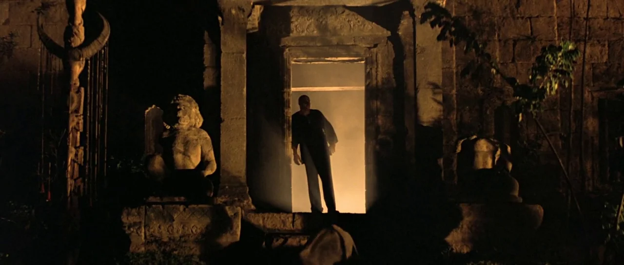 Apocalypse Now (1979) movie still: wide — A stone structure features ornate carvings and statues flanking a central dark entranceway. A man stands…