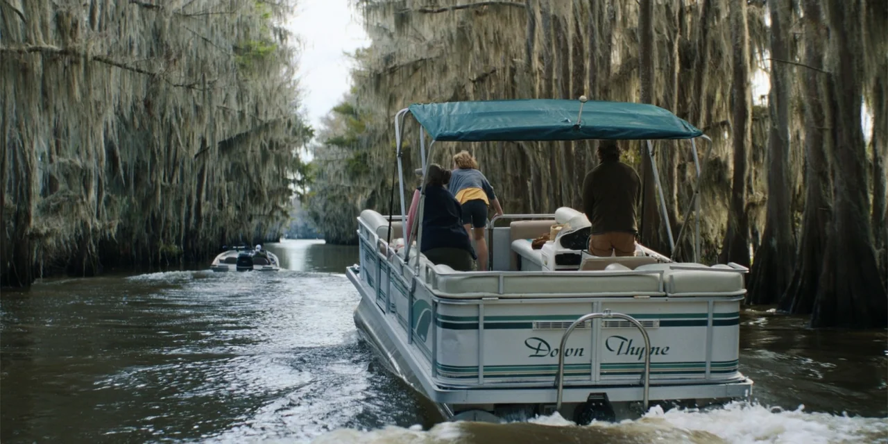 Caddo Lake (2024) movie still: wide — A pontoon boat navigates through a narrow channel surrounded by dense cypress trees draped in moss. Three…