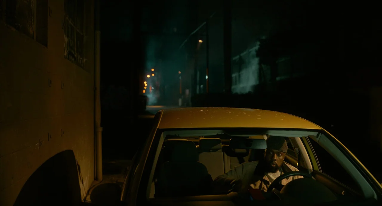 Fresh (2022) movie still: medium — A man sits in the driver seat of a parked car on a narrow street. Dark urban surroundings feature distant street…