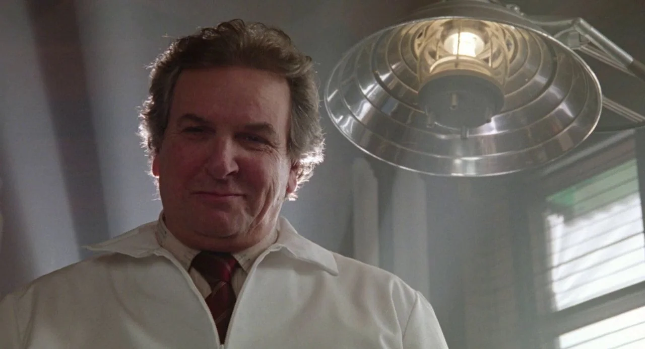 Jacob’s Ladder (1990) movie still: closeup — A clinical space is illuminated by a single high-intensity metal desk lamp overhead. A middle-aged man…