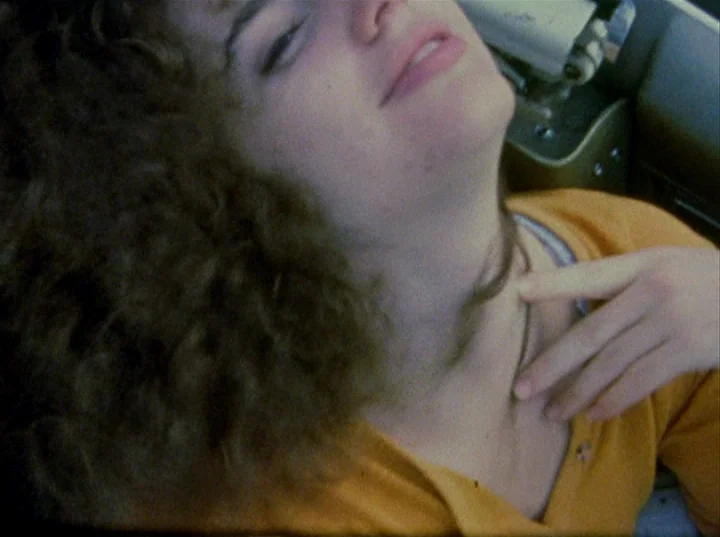 Slacker (1991) movie still: closeup — The interior of a 1970s vehicle shows dashboard elements in the blurry background. A young woman with curly…