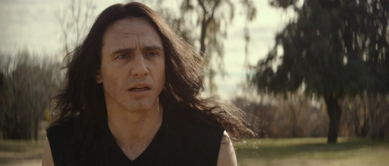 The Disaster Artist (2017) movie still: closeup — An outdoor park setting features blurred trees and grass under a bright daylight sky. A man with…