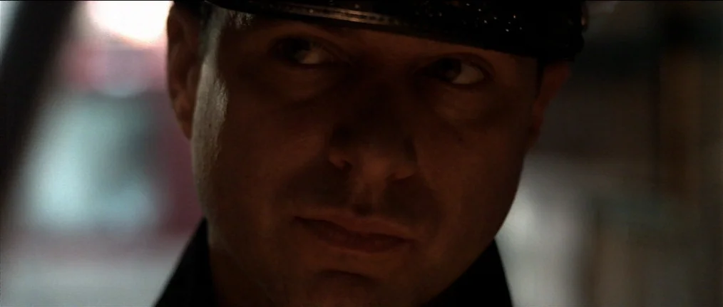 Se7en (1995) movie still: closeup — A dark, dimly lit indoor space with shadows obscuring most of the background. A man wearing a dark peaked hat…