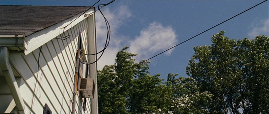 Take Shelter (2011) movie still: establishing — A low-angle view shows the white siding and dark roof of a suburban house. Electrical wires cross…