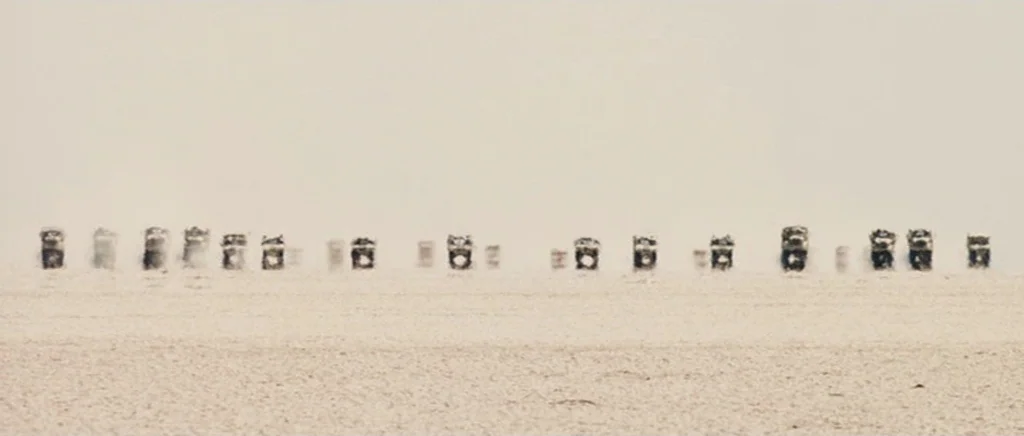 Jarhead movie still: wide — A vast, flat, light-colored expanse stretches toward a hazy, monochromatic horizon under bright light. A long line of…